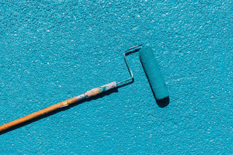 A close-up of a blue paint roller applying paint on a textured wall surface, captured in bright sunlight.